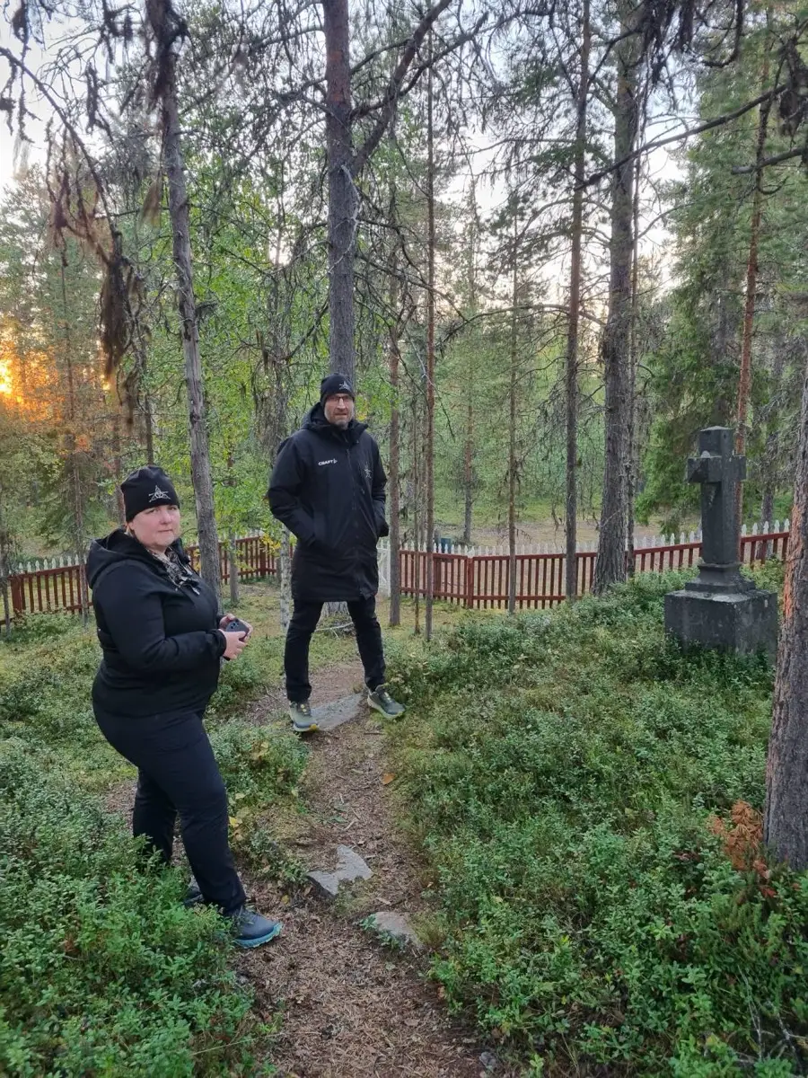 Två personer i svarta kläder står vid en inhägnad skogskyrkogård med ett stenkors, i kvällssol