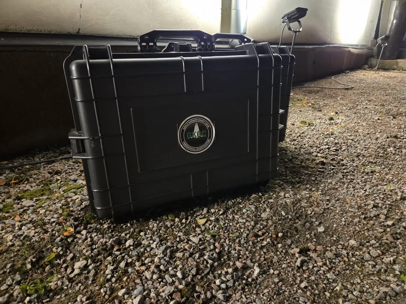 Ett svart flightcase med GKGC:s emblem står på grus under en byggnad