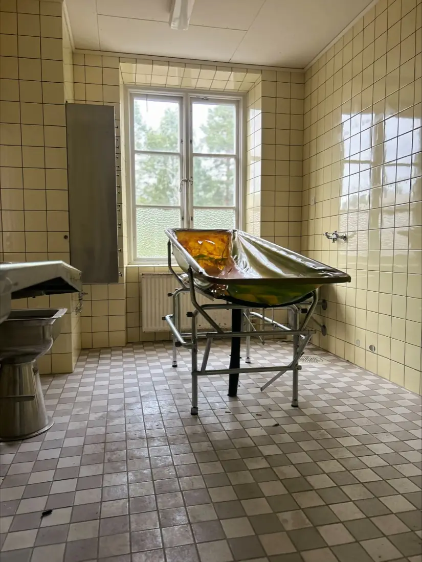 Et flisebeklædt behandlingsrum med en gammel badebåre på stativ